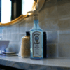 Turquoise bombay sapphire soap dispenser with vintage gold pump placed on a bathroom vanity made of marble. Next to it is a bottle of gold bath salts, a white sink and some coasters. Above it is a gold-rimmed circular mirror