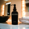 Bulldog soap dispenser with matte black pump placed in between a metallic bathroom sink and set of white fluffy towels rolled up like in a hotel