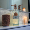 Tribute soap dispenser placed on bathroom vanity next to a toothbrush holder. On the right is a lit candle and behind it is a mirror